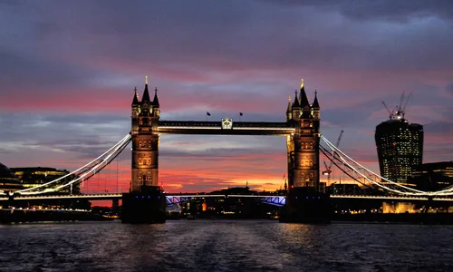 This urban landscape photograph captures London’s iconic Tower Bridge at sunset during the evening in early autumn. The illuminated architecture of the bridge stands out against the vibrant sky, which is painted with shades of orange, red, and purple as the sun sets over the city. The image showcases one of the United Kingdom’s most famous bridges and highlights the unique blend of historical and modern design characteristic of London. City lights begin to sparkle around the urban waterfront, further emphasizing the architectural features of Tower Bridge in this prominent city setting.